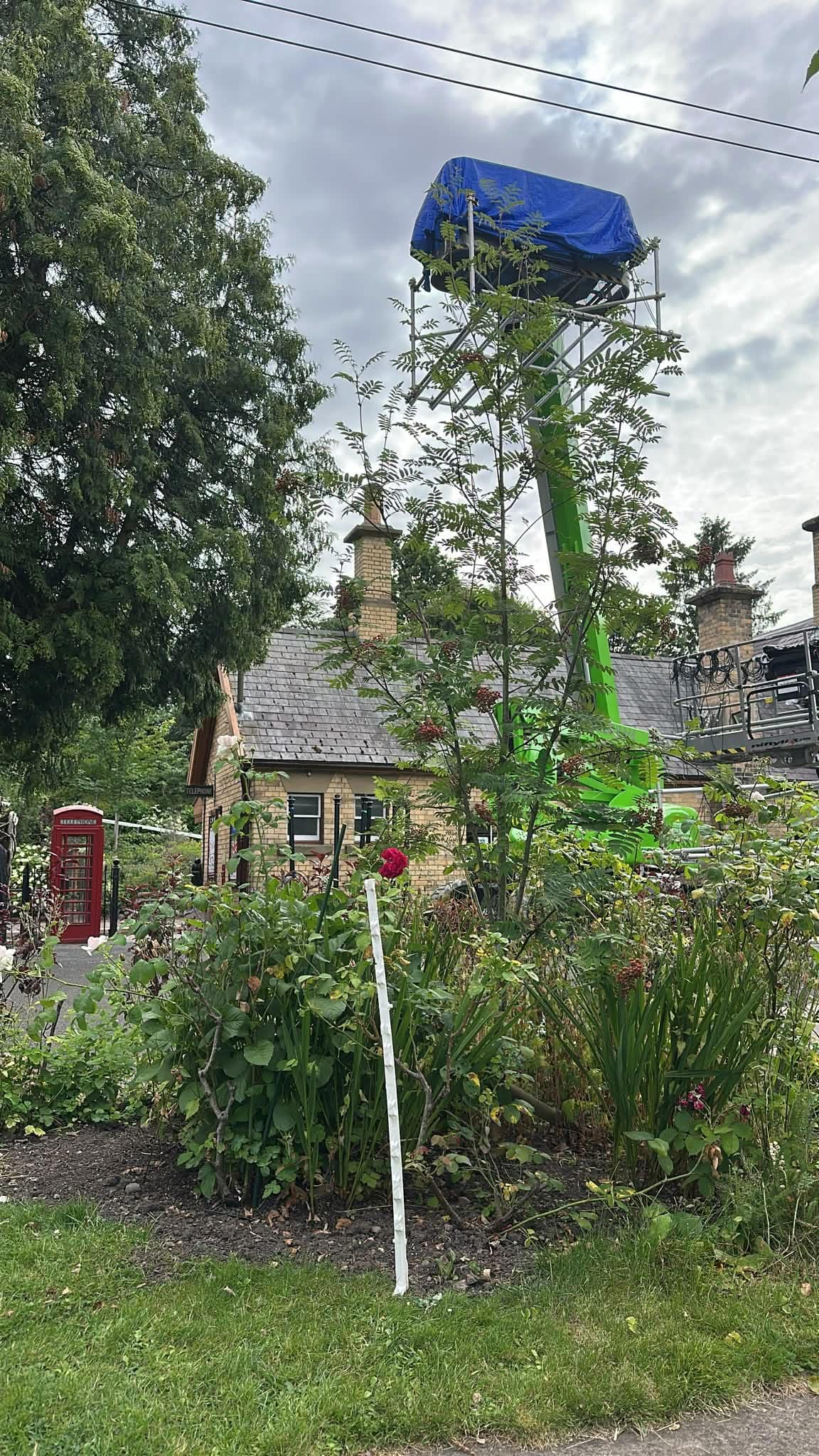 Green cherry picker rigging lighting equipment above a period cottage on a rural film location.